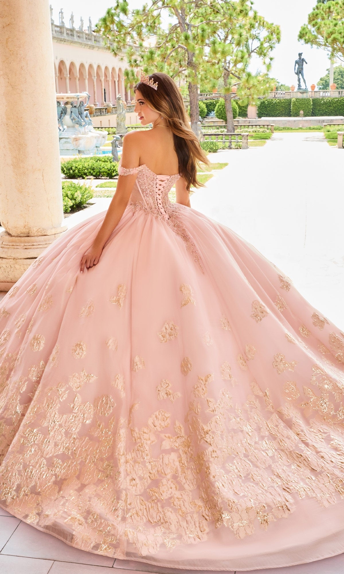 A woman stands outdoors near columns, facing away, wearing the Quinceanera Dress PR30154 by Princesa—a pink ball gown with gold floral details and a sweetheart neckline.